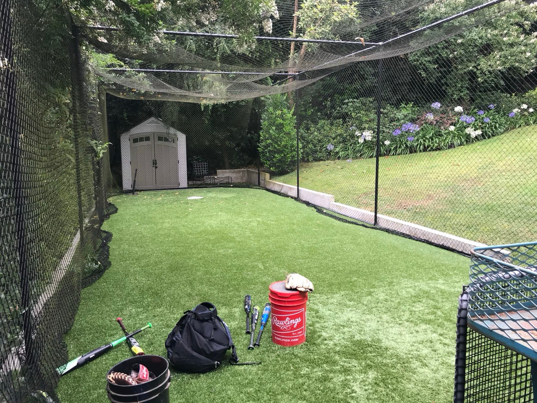 Backyard batting cage with turf, a storage shed, a bucket of baseballs and bats on the ground